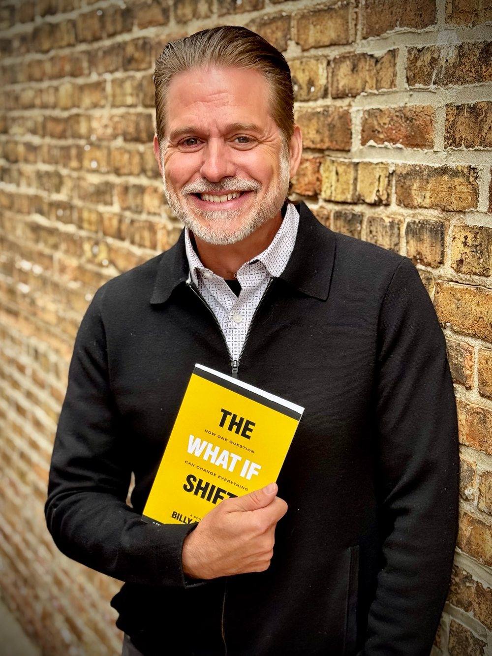 Billy Hyland holding a copy of The What If Shift, smiling against a brick wall